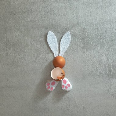 Şirin tavşancık beyaz kulaklar, tüylü pembe pençeler ve gri beton arka planda yumurta kabukları. Metin uzayı. Üst Manzara. Asgari biçim.
