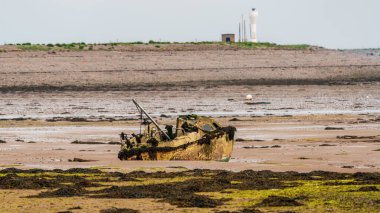 Walney Kanalı 'nın çamurunda hasar görmüş bir tekne, arka planda bir deniz feneriyle Roa Adası, Cumbria, İngiltere' ye giderken görülmüş.