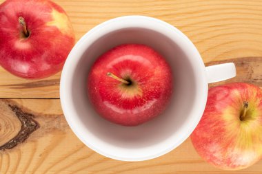 Tahta bir masanın üstünde beyaz seramik bardakla üç tatlı organik, kırmızı elma..