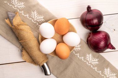 Seramik bir tabakta keten peçeteyle bej ve beyaz organik tavuk yumurtası. Yakınındaki metal bıçakta jüt peçete ve beyaz boyalı doğal ahşaptan yapılmış bir masada iki soğan var..