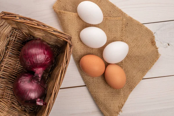 Birkaç taze organik lezzetli, bej ve beyaz, jüt peçeteli tavuk yumurtası ve beyaz ahşap tezgahın üstünde sepette iki soğan..