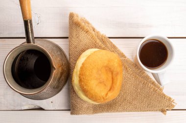 Bir tane taze pişmiş iştah açıcı, lezzetli, kirazlı tatlı çörek jüt peçeteye bir fincan kahve ve yakın plan, ahşaptan boyanmış beyaz bir masa, üst görünüm.