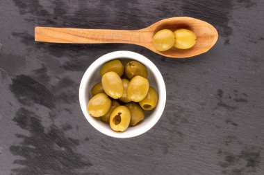 Birkaç baharatlı organik turşulu yeşil zeytin, beyaz bir seramik tabağın üzerinde, arka planda tahta bir kaşık, en üstteki manzaralı..