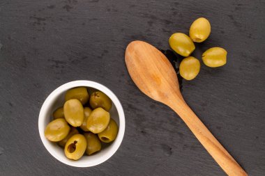 Birkaç baharatlı organik turşulu yeşil zeytin, beyaz bir seramik tabağın üzerinde, arka planda tahta bir kaşık, en üstteki manzaralı..