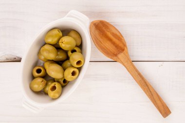 Birkaç baharatlı organik turşulu yeşil zeytin, beyaz bir seramik tabağın ve ahşap bir kaşığın üzerinde uzanır..