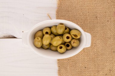 Birkaç baharatlı organik turşulu yeşil zeytin turşusu beyaz seramik bir tabağın üzerinde duruyor. Beyaz, doğal ahşap bir zemin üzerinde jüt bir peçete..