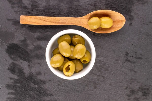 Birkaç baharatlı organik turşulu yeşil zeytin, beyaz bir seramik tabağın üzerinde, arka planda tahta bir kaşık, en üstteki manzaralı..