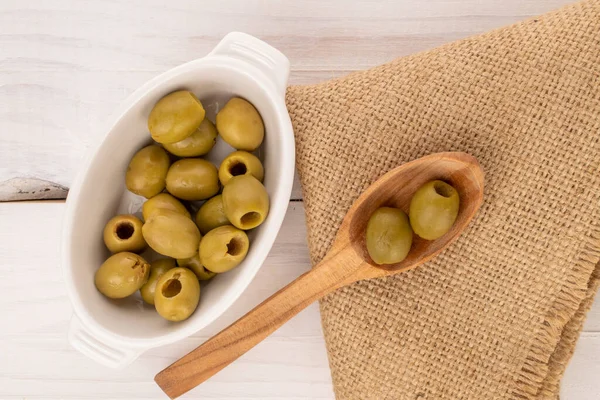 Birkaç baharatlı organik turşulu yeşil zeytin turşusu beyaz bir seramik tabağın üzerinde, tahta bir kaşık ve arka planda bir jüt peçete, beyaz boyalı ahşap, üst manzara.