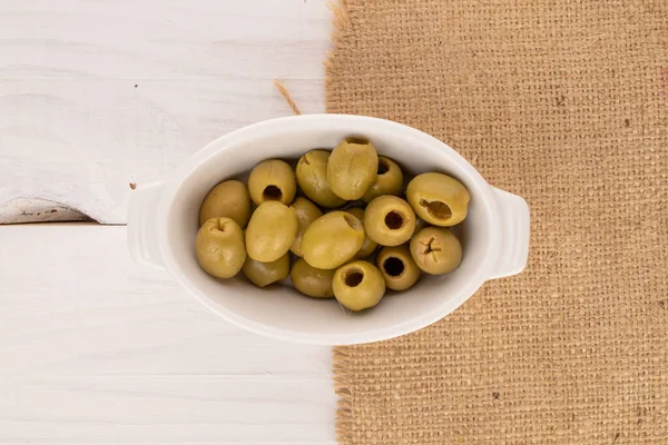 Birkaç baharatlı organik turşulu yeşil zeytin turşusu beyaz seramik bir tabağın üzerinde duruyor. Beyaz, doğal ahşap bir zemin üzerinde jüt bir peçete..