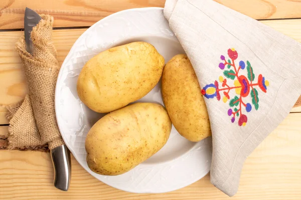 三个新鲜的 没有剥皮的 有机多汁的土豆 用金属刀包在黄麻餐巾上 在陶瓷白盘上和亚麻布餐巾上 在天然的木桌上 — 图库照片