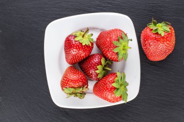 Birkaç olgun organik çilek, beyaz bir seramik çay tabağı ile kayrak taşı üzerinde, yakın plan, üst manzara..