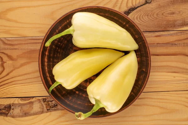 Three bright yellow sweet peppers with a clay dish on a wooden table, close-up, top view.