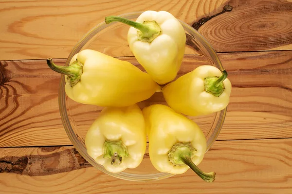 Several bright yellow sweet peppers in a glass bowl on a wooden table, close-up, top view.