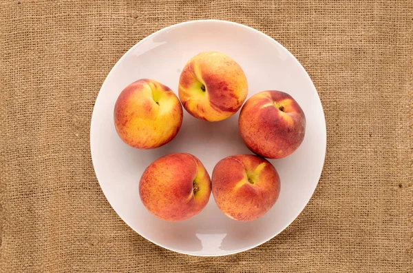 Birkaç sulu olgun şeftali, beyaz bir seramik tabakla jüt kumaş üzerinde, yakın plan, üst manzara..