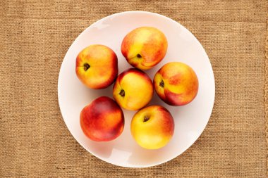 Jüt kumaşında beyaz seramik bir plaka ile birkaç olgun sulu nektarin, yakın plan, üst görünüm.