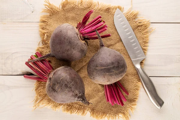 Three sweet red beetroots with a jute napkin and a metal knife on a wooden table, close-up, top view.