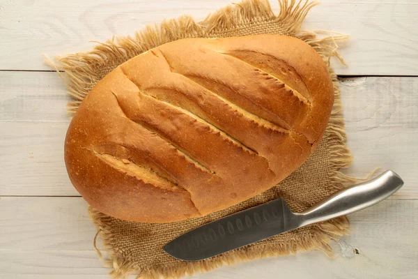 One fragrant fresh loaf with a jute napkin and a knife on a wooden table, close-up, top view.