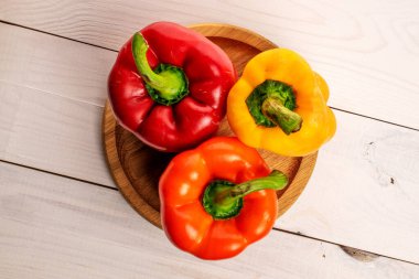 Three whole organic fresh juicy tasty bell peppers, close-up, on a round bamboo tray, standing on a painted white tabletop made of wood, top view.