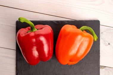 Two whole organic fresh juicy tasty bell peppers, close-up, on a serving plate of slate lying on a painted white tabletop made of wood, top view.