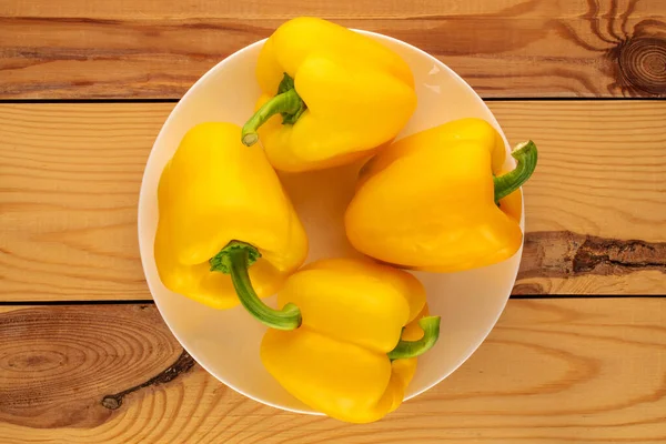 Four ripe yellow sweet peppers with a ceramic plate on a wooden table, close-up, top view.