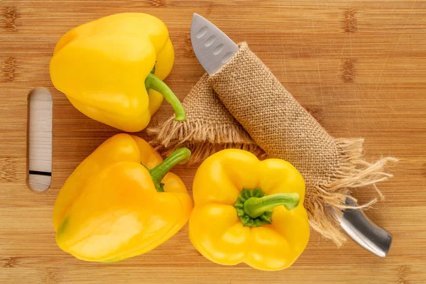 Three ripe yellow sweet peppers with a bamboo cutting board and a knife on a wooden table, close-up, top view.