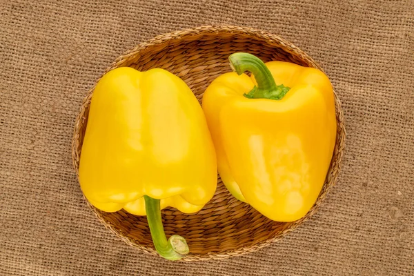 Two ripe yellow sweet peppers in a straw plate on a jute cloth, close-up, top view.