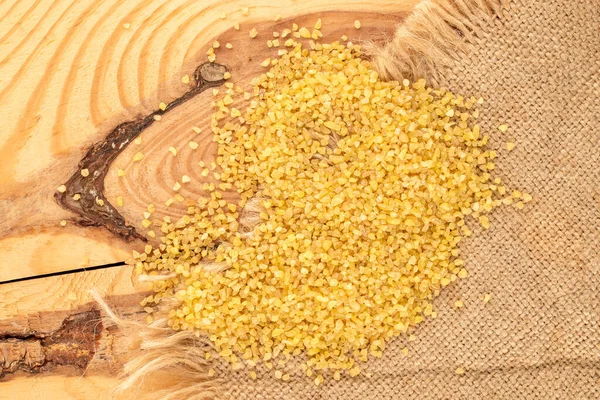 Raw bulgur grains with jute napkin on wooden table, macro, top view.