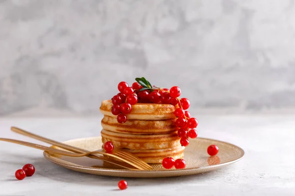 Stack of healthy oat pancakes with red currant in a golden plate with golden cutlery on grey stone background horizontal