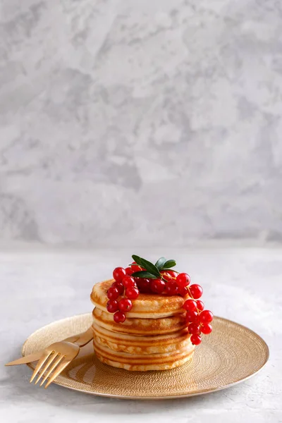 Stack of healthy oat pancakes with red currant in a golden plate with golden cutlery on grey stone background vertical