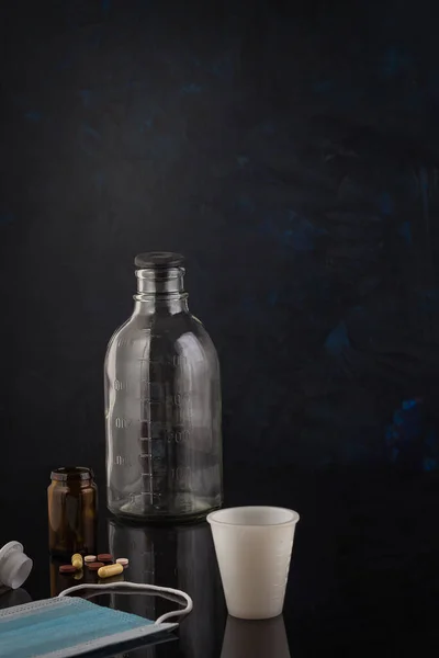 Siyah camda peş peşe ilaç şişeleri, haplar, tıbbi maske, ölçüm bardağı. Virüs tedavisi konsepti. Uzayı kopyala, odağını kaldır