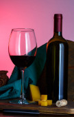 sklenice a láhev červeného vína s tvrdým sýrem na růžovém pozadí, rustikální styl. Sada červeného vína na dřevěné desce, zátiší, selektivní ostření, kopírovací prostor.