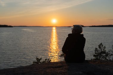 Bir kadın kayalıklarda, deniz kıyısında oturur, adam gün batımını izler. Dinlenme konsepti, tatil.