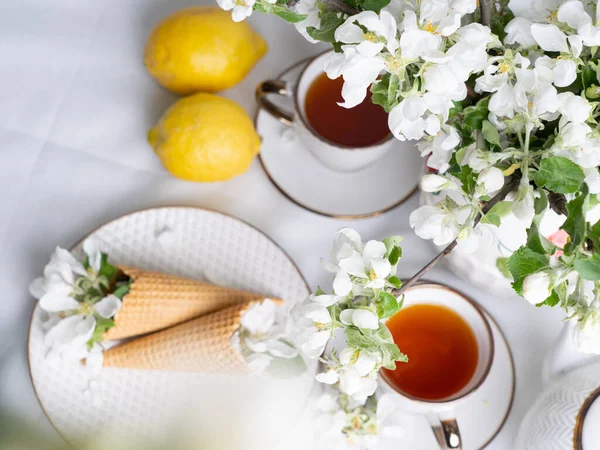 White tea set with tea in cups with yellow lemons and a blossoming apple branch.View from aboveThere are waffle cones on a saucer.