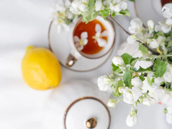 White tea set with tea in cups with yellow lemons and a blossoming apple branch.Close-upThere are waffle cones on a saucer.