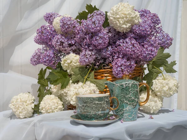 A bouquet of lilacs, white viburnum in a basket and a tea set. on the table in the garden, the background is white