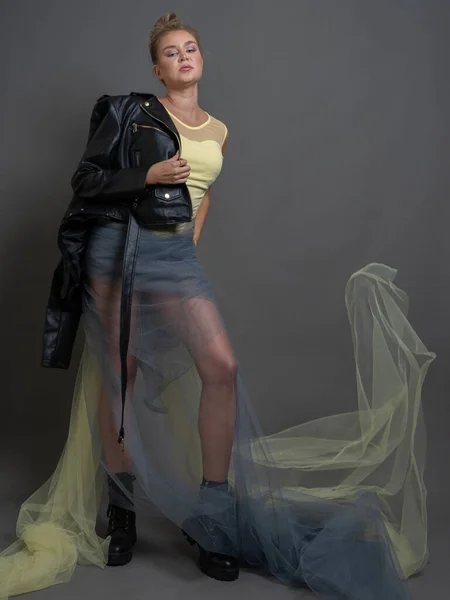 Fashion blonde woman in black eco leather boots, jacket, transparent tulle skirt, studio shot on gray background, vertical photo, full length portrait