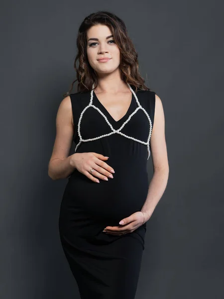 Fashionable pregnant mom hugs her belly. Stylish motherhood concept. Happy young pregnant woman in stylish black dress and pearl accessory. Medium shot portrait against a dark background.