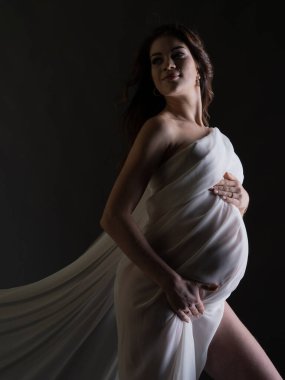 Güzel beyaz ipek kumaşlı hamile bir kadının portresi. Gri bir arka planda dikey fotoğraf. Tek ışık kaynağıyla stüdyo fotoğrafı.