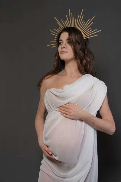 Portrait of a beautiful pregnant woman in a crown, like the rays of the sun, and with a beautiful white silk fabric. Vertical photo on a gray background.