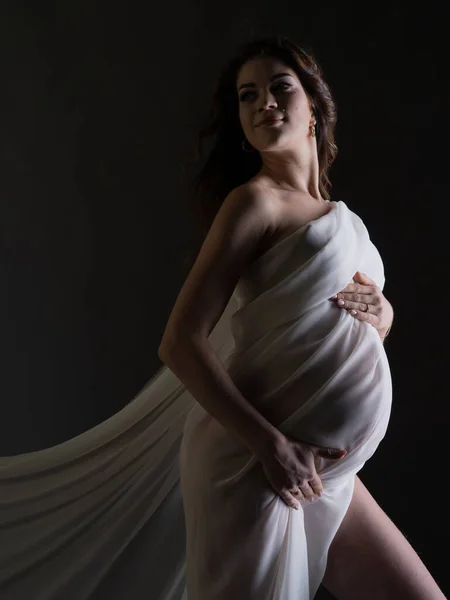 Güzel beyaz ipek kumaşlı hamile bir kadının portresi. Gri bir arka planda dikey fotoğraf. Tek ışık kaynağıyla stüdyo fotoğrafı.
