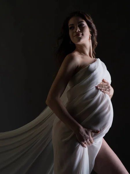 一个美丽的怀孕妇女的画像 她有美丽的白色丝织品 灰色背景上的垂直照片 一个光源的演播室照片 图库照片