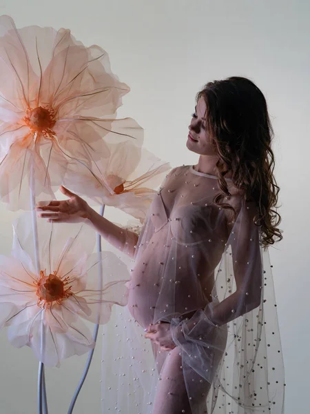 ヌードランジェリーの若い妊婦の公開写真や真珠で飾られた透明なドレス 光の背景と完全な成長で巨大な装飾的な花と — ストック写真