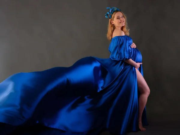 A pregnant girl with blonde hair in a long blue dress with a flowing train and a headband with butterflies poses in front of the camera. The concept of a happy pregnancy