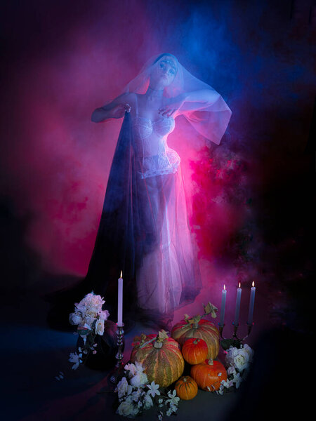 Portrait of a mysterious attractive woman. Red background, white lace top, skirt and white veil on the head. A beautiful passionate girl is holding a huge black cloth in her hands, and candles are burning around. The concept of a Halloween party.