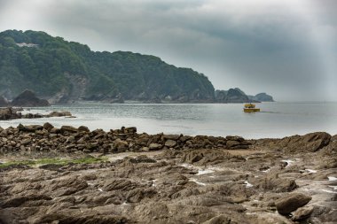 Bir deniz koyundaki kayalık sahili ve uzaktaki kayalıkları gösteren küçük bir teknenin manzarası.