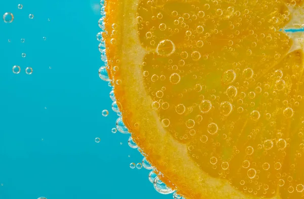 Parlak mavi arkaplanda kabarcıklar gösteren sıvı içinde asılı duran portakal dilimini kapat