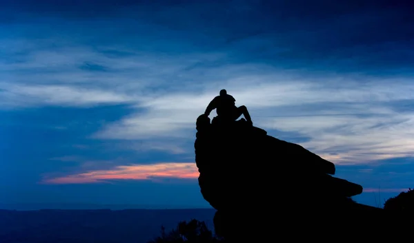 Tanımlanamayan bir adam gölgede kayanın üzerinde oturuyor. Arkasında koyu mavi ve pembe bir akşam gökyüzü var.