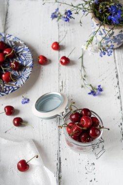 Cam kavanozda tatlı kirazlar ve beyaz ahşap arka planda mavi çiçekli bir tabak.
