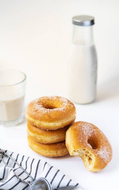 Üç koca çörek ve bir tanesi beyaz ahşap masada, süt şişesi, cam elek ve peçeteyle ısırılıyor.
