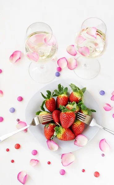 Концепция Дня Святого Валентина. Игристое вино в двух бокалах, клубника в тарелке с двумя вилами, лепестки роз — стоковое фото